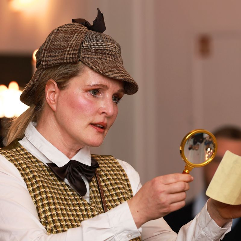 Eliza mit Sherlock Hut und Weste und Lupe untersucht Papierkarte