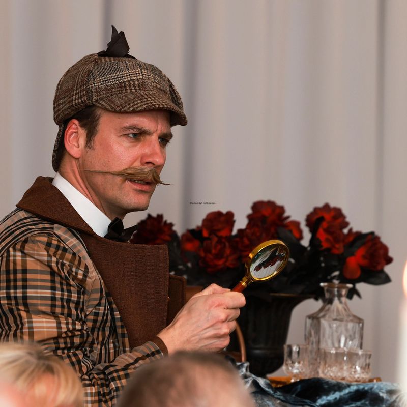 Conan Doyle im Sherlock Holmes Look mit Lupe vor Stehtisch mit Rosenvase und Kerzen